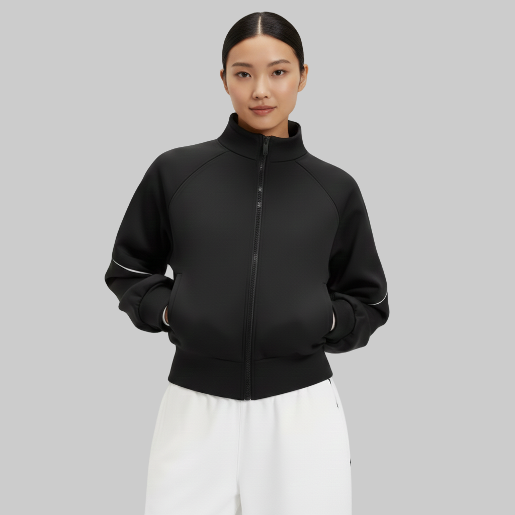 Kobieta ubrana w czarną kurtkę z zamkiem błyskawicznym i wysokim kołnierzem o gładkim, dopasowanym kroju, z rękami w kieszeniach, pokazana od przodu na szarym tle.