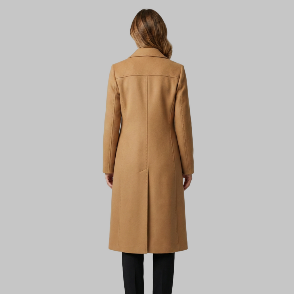 Kobieta w brązowym płaszczu i białym swetrze z golfem stoi pewnie z rękami w kieszeniach. Ma na sobie czarne spodnie i uśmiecha się na szarym tle.