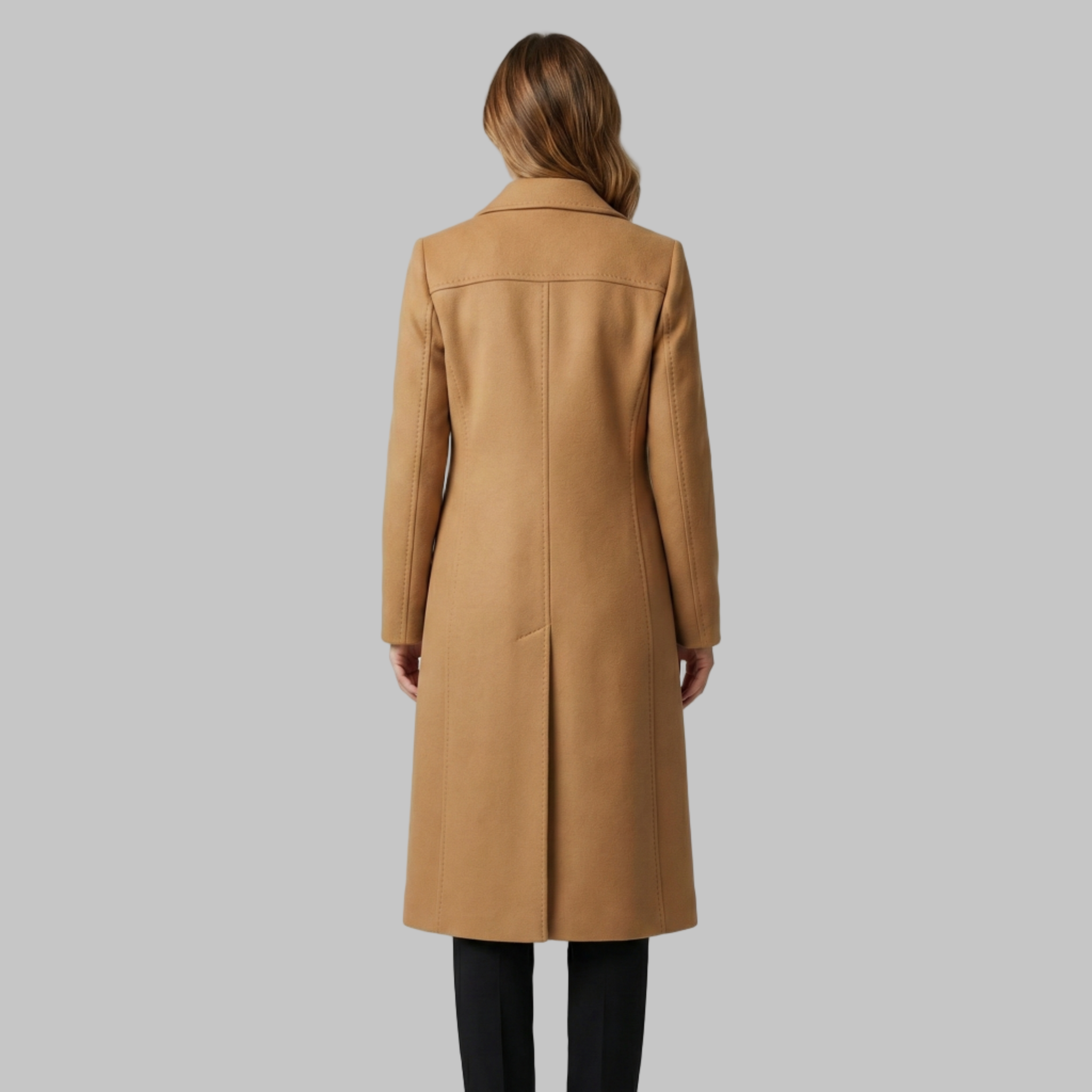 Kobieta w brązowym płaszczu i białym swetrze z golfem stoi pewnie z rękami w kieszeniach. Ma na sobie czarne spodnie i uśmiecha się na szarym tle.