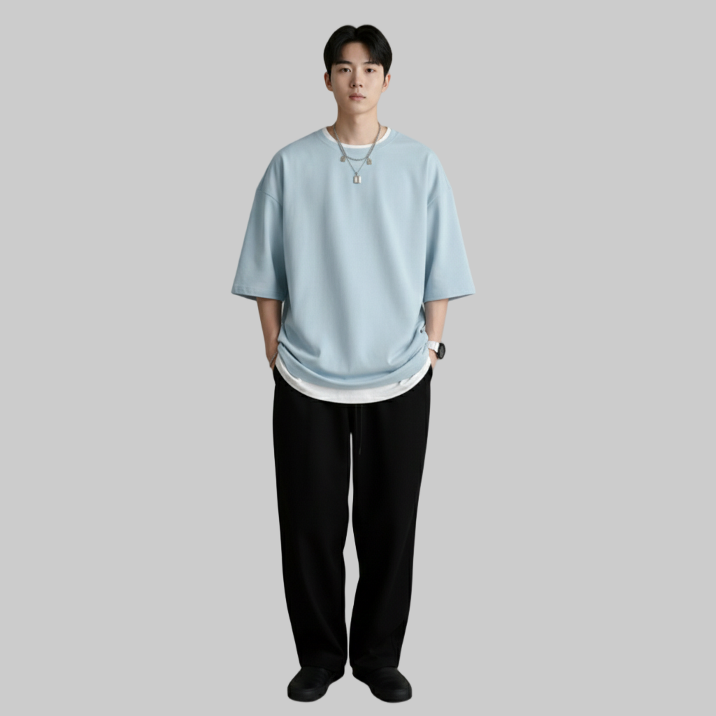 Mężczyzna ubrany w oversize'ową jasnoniebieską koszulkę z białym obszyciem, czarne luźne spodnie i czarne buty, stojący na szarym tle.