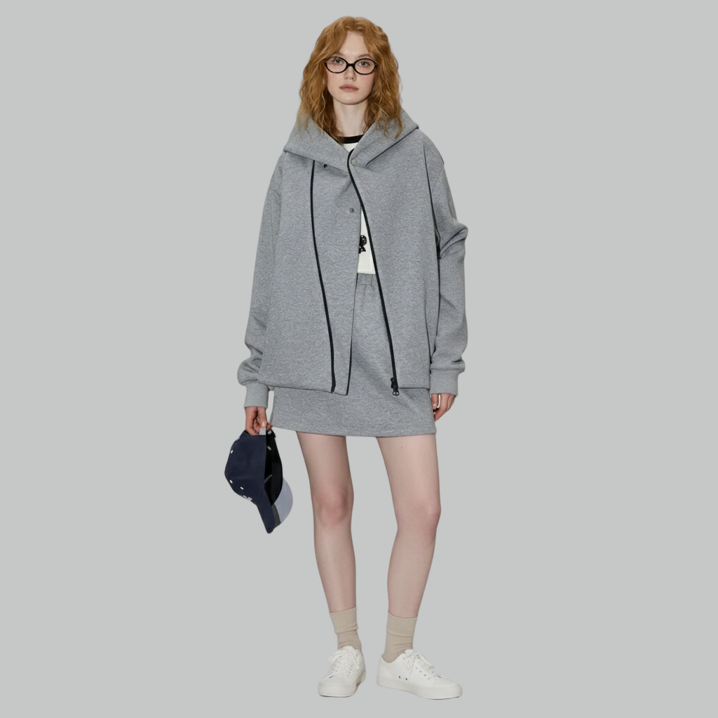 Szary zestaw damski składający się z oversize'owej bluzy z kapturem zapinanej na zamek błyskawiczny i dopasowanej mini spódniczki, stylizowany z białymi trampkami i beżowymi skarpetkami.