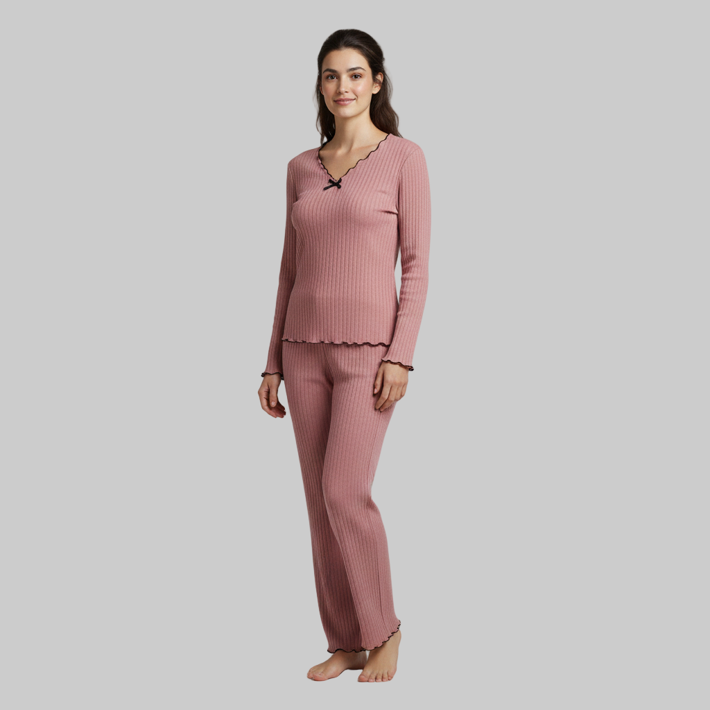 Kobieta ubrana w różowy, prążkowany zestaw domowy z długimi rękawami i czarnym wykończeniem w kształcie muszelki, stojąca boso na szarym tle.