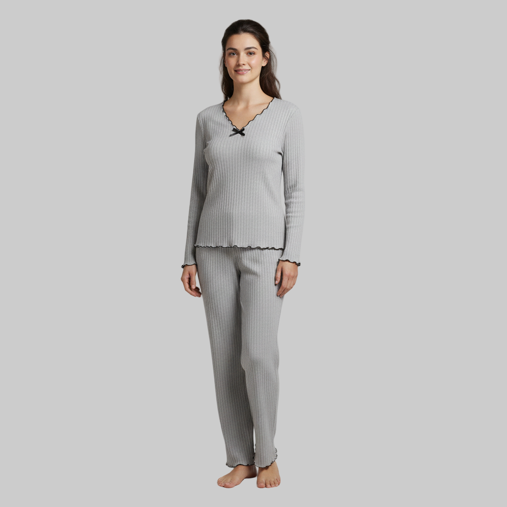 Kobieta ubrana w jasnoszary zestaw domowy z długimi rękawami i czarnymi falbankami, stojąca boso na szarym tle.